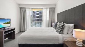 Hotellihuone, jossa on suuri sänky, puinen TV-taso, yöpöytälamppu ja ikkunasta avautuva näkymä korkeaan kerrostaloon.