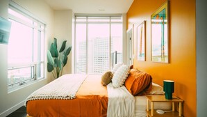 A bedroom with a large window, a bed with orange bedding, a bedside table with a lamp, and artwork on the wall. - *Free Parking* Cozy 2br/2ba Penthouse DT Halifax! (Halifax)