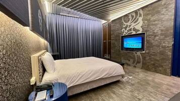 A bedroom with a bed, a flat-screen TV mounted on the wall, a bedside table with a lamp, and a curtain.
