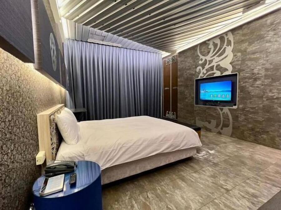 Una habitación de hotel con una cama, un televisor montado en la pared, una mesita auxiliar azul y un papel tapiz estampado.