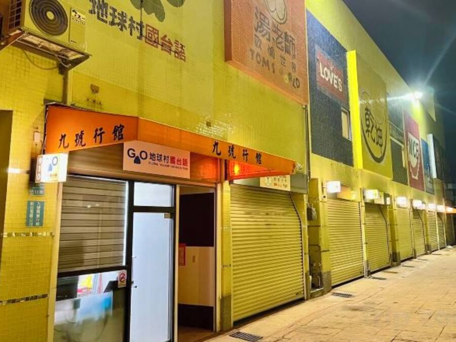 A building facade with multiple closed shuttered storefronts, a visible entrance, and signage in a foreign language.