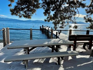 Mesa e banco de piquenique cobertos de neve à beira de um lago, com um cais que se estende sobre a água.