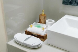 A bathroom with a white sink, a wooden soap dispenser, a roll of toilet paper, a stack of white towels, and a small wooden box with a towel and a flower. - Moning Villa Ubud (Bali)