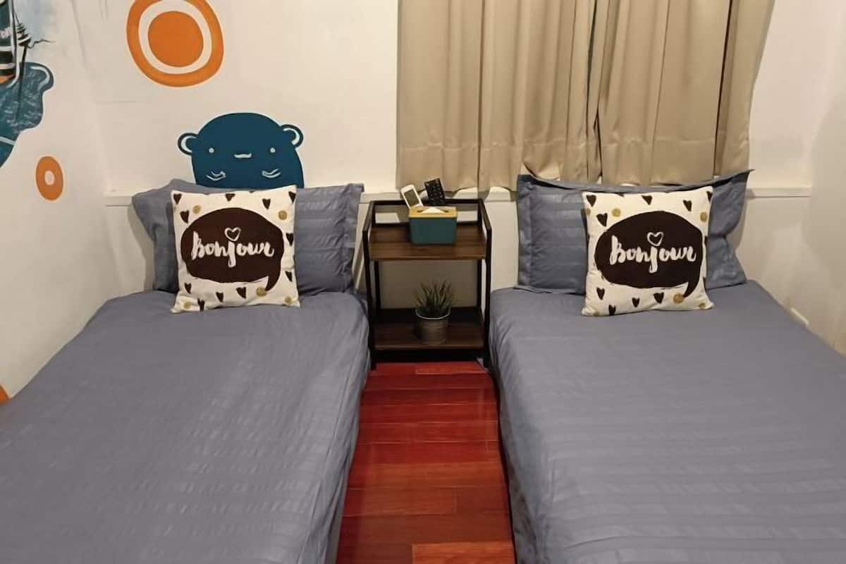 Una habitació amb dos llits, cadascun amb un coixí que mostra la paraula 'bonjour' i un disseny d'ós. Hi ha una taula petita amb una planta i una caixa entre els llits.