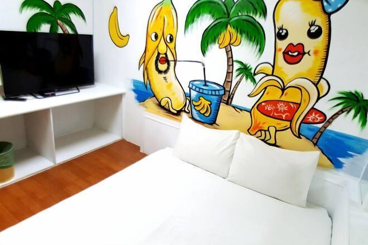 Habitació amb llit blanc, televisió de pantalla plana sobre un suport i un mural de plàtans a la paret.