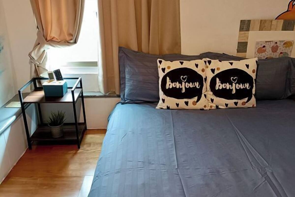 Un dormitori amb un llit, dos sofàs d'abraçar amb el text 'Bonjour', una taula de nit amb una làmpada, una planta i una finestra amb cortines.