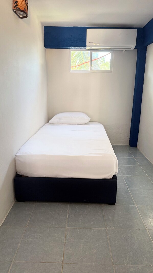 Une petite chambre avec un lit simple, un matelas blanc, une base de couleur sombre, une fenêtre donnant sur des palmiers et un climatiseur fixé au plafond.