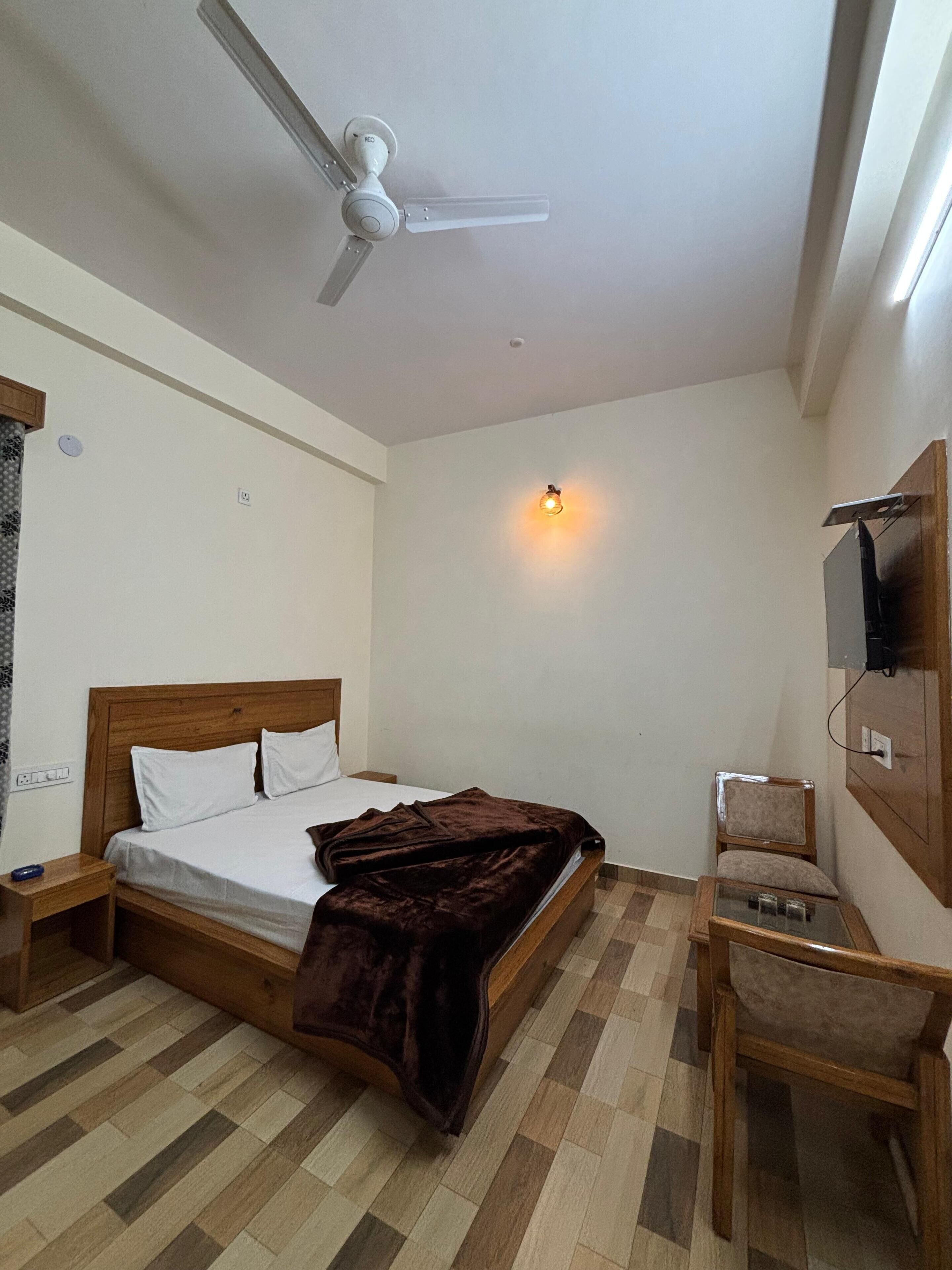 Un dormitorio con cama, mesita de noche de madera, silla, escritorio, ventilador de techo, televisor montado en la pared y luz adosada a la pared.