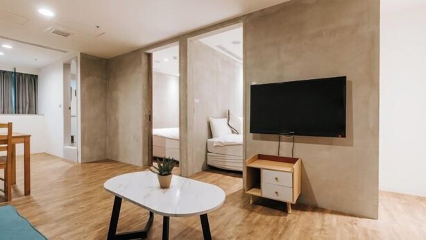 Een moderne hotelkamer met een houten vloer, een witte ronde tafel met een plant erop, een bed met wit beddengoed, een aan de muur bevestigde televisie, en een houten kast.