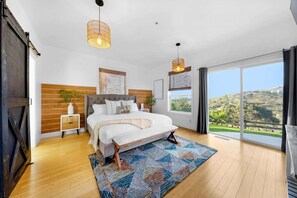 A bedroom with a large bed, wooden panel wall, two pendant lights, a view of greenery through a large window, and a sliding door.