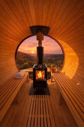 A circular wooden room with a wood-burning stove, benches, and a view of the landscape.