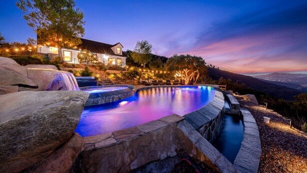 A swimming pool with a waterfall feature, surrounded by stone walls and illuminated by ambient lighting. - San Diego Serenity Hills, Pickleball, Golf, Infinity Pool with Epic Views (Jamul)
