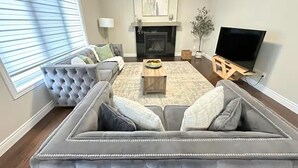 Una sala de estar con un amplio sofá modular, una chimenea, un televisor y un ventanal con persianas.