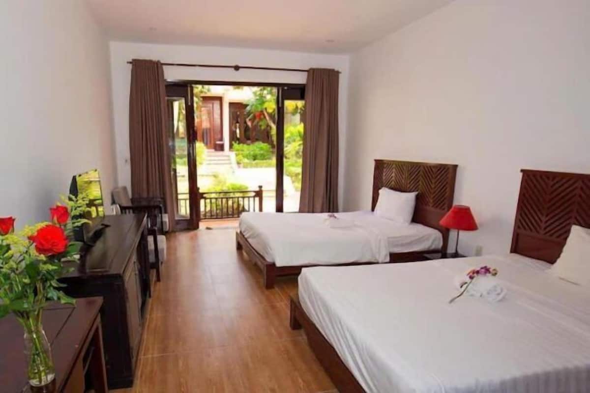 A hotel room with two beds, a desk, a chair, a vase with flowers, and a view of the outdoors.
