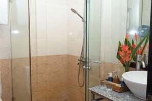 Een douchecabine met glazen wanden, een handdouche, een wastafel met een marmeren blad en een vaas met bloemen.