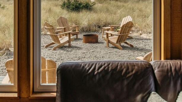 A room with a large window, wooden paneled walls, and a view of a grassy landscape with outdoor seating. - Lighthouse on the Beach with Hot Tub & Fire Pit (Manzanita)