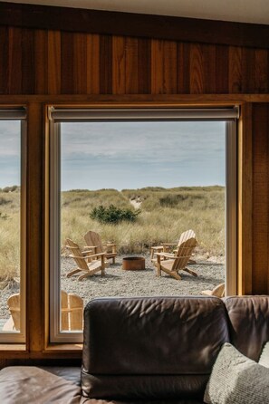 A room with a large window, wooden paneled walls, and a view of a grassy landscape with outdoor seating. - Lighthouse on the Beach with Hot Tub & Fire Pit (Manzanita)