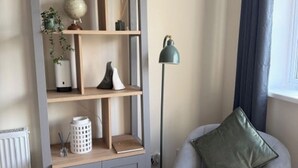 Huoneessa on harmaa sohva, vihreä koristetyyny, lattiavalaisin ja puinen hylly, jonka päällä on koriste-esineitä.