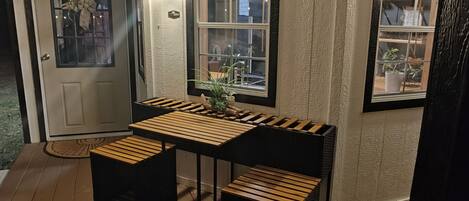 Un espace extérieur aménagé avec des meubles en bois, comprenant une table et des bancs, une porte ornée d’une pancarte indiquant « Soyez les bienvenus », et de grandes fenêtres derrière lesquelles on aperçoit des plantes.