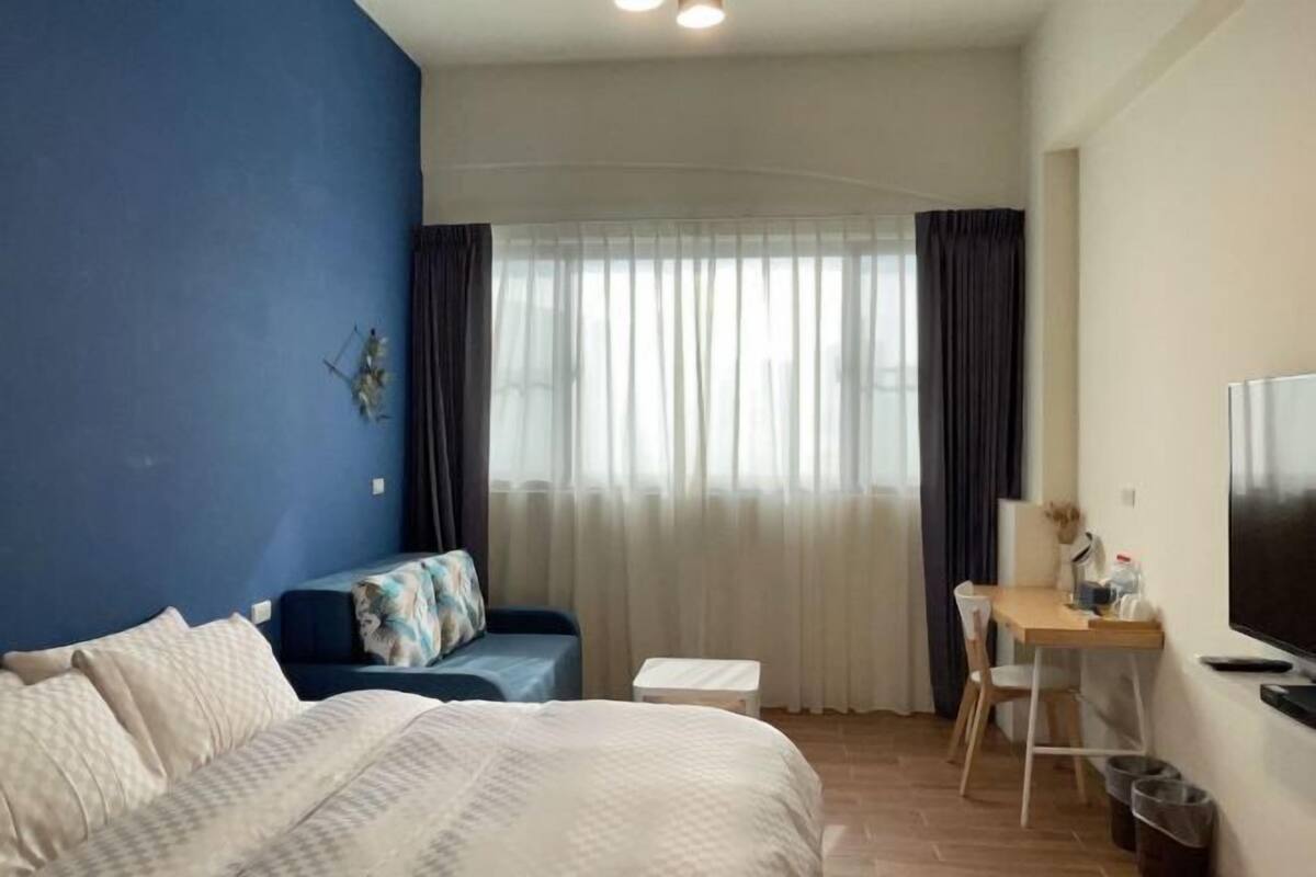 A bedroom with a bed, a desk, a chair, a TV, and a window with curtains.