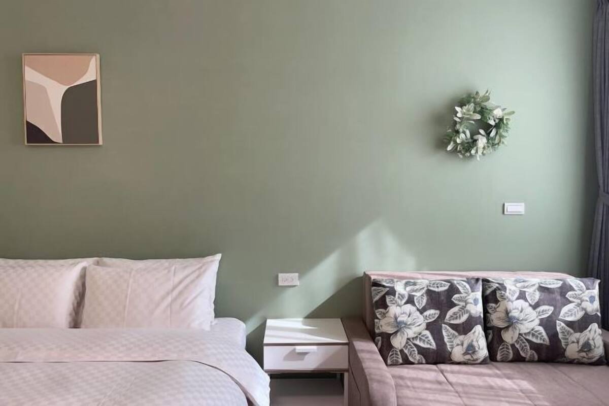 A bedroom with a bed, a nightstand, a sofa with floral cushions, a wall-mounted artwork, and a small plant.