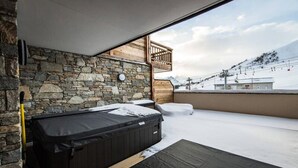 Habitación con pared de piedra, banca de madera, bañera de hidromasaje y vistas a una montaña nevada y una pista de esquí.