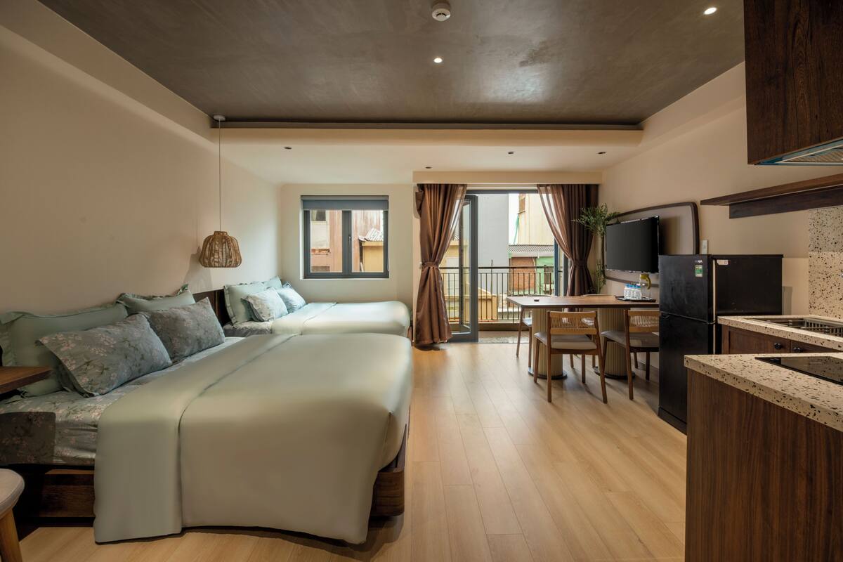 A modern bedroom with a large bed, a small desk, and a kitchen area with a refrigerator and a TV.