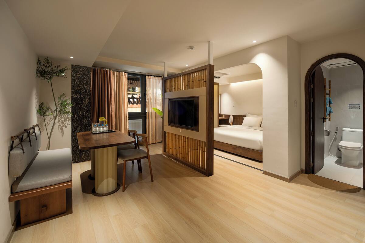 A modern hotel room with a wooden floor, a sofa, a TV, and a bathroom visible through an open door.