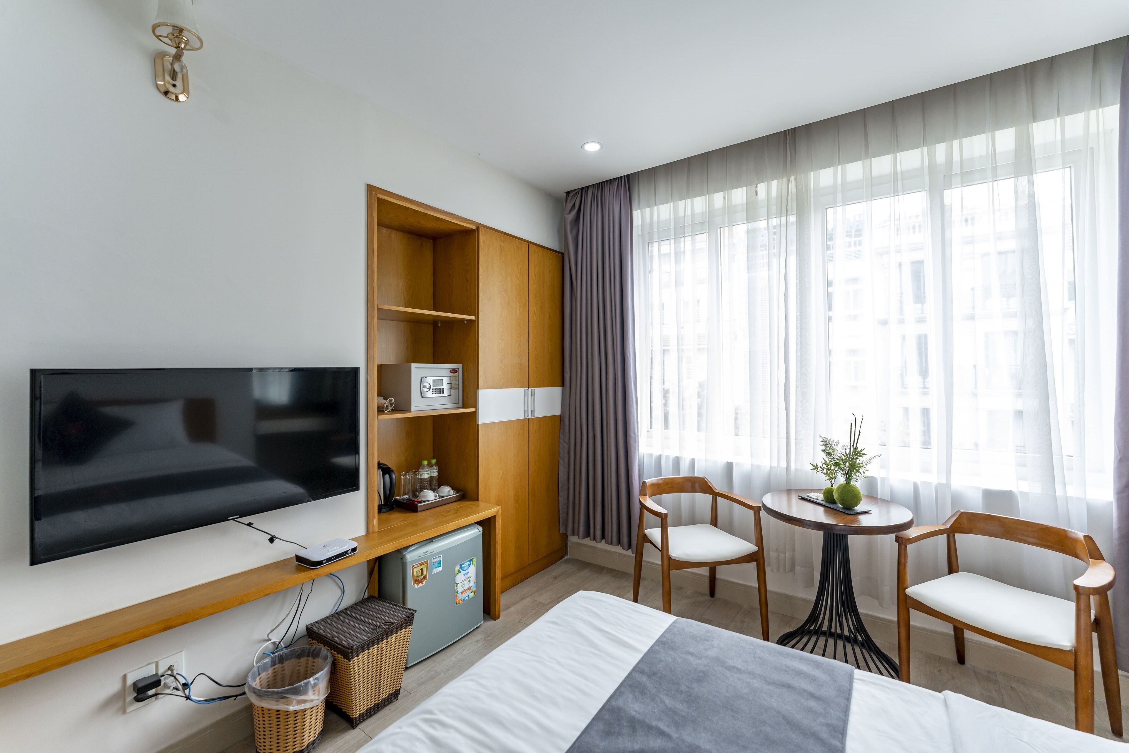 A modern hotel room with a flat-screen TV, wooden furniture, a small refrigerator, wicker baskets, and a large window with curtains.