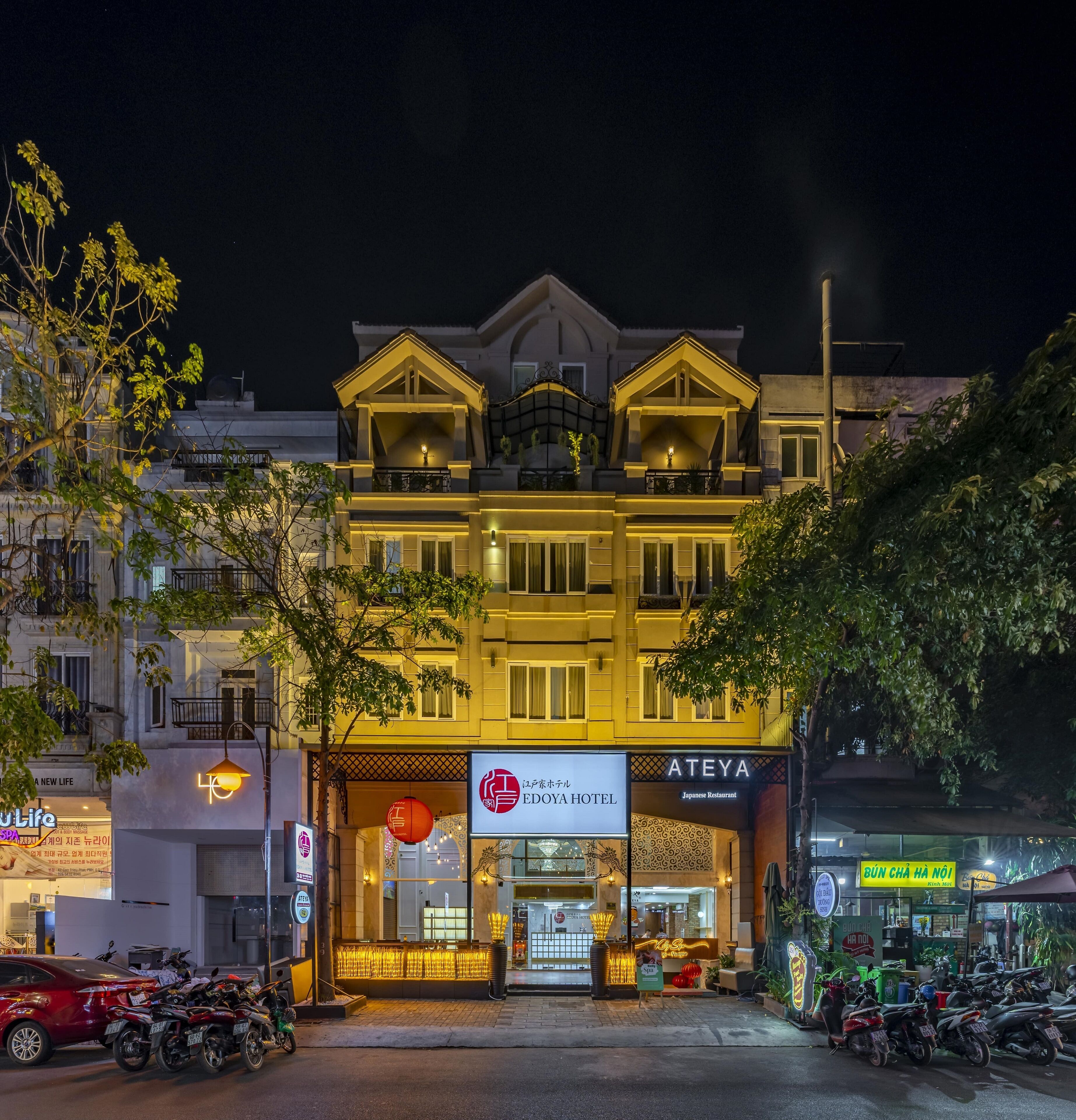 A hotel with a traditional architectural style, featuring a prominent entrance with a signboard, a well-lit facade, and a parking area with motorcycles.