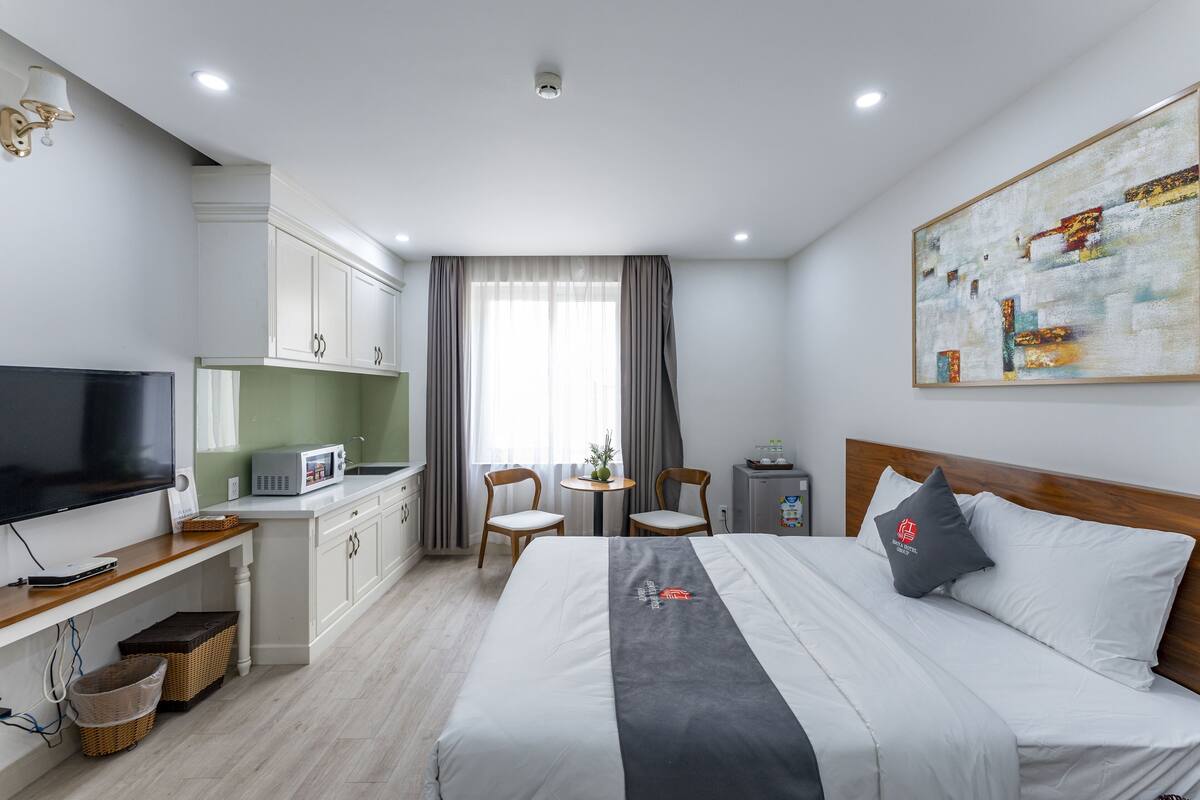 A modern hotel room with a bed, a TV, a kitchenette, and a dining area.