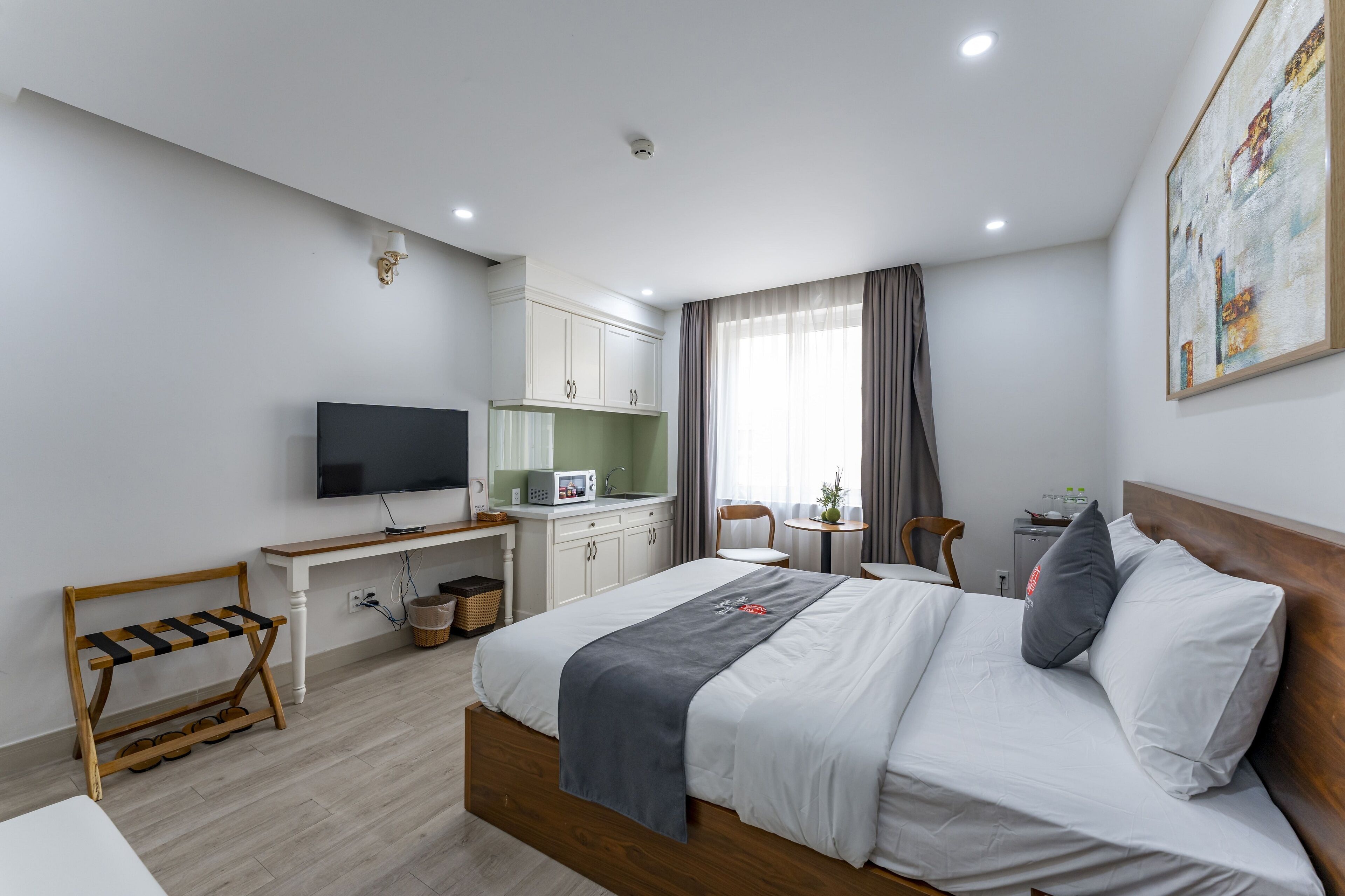 Une chambre d’hôtel moderne équipée d’un lit, d’un bureau avec un téléviseur à écran plat, d’une petite kitchenette et d’une fenêtre avec des rideaux.
