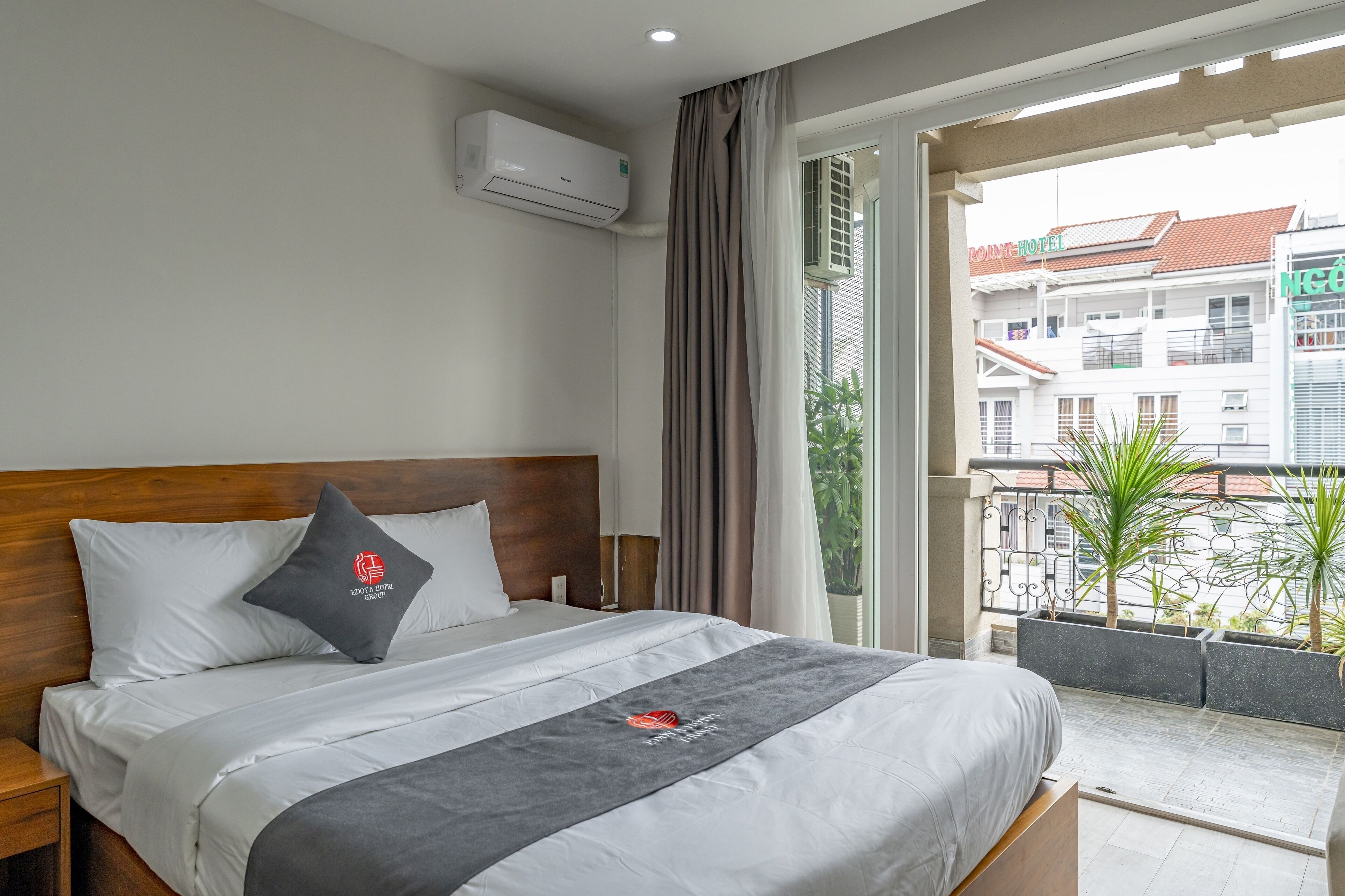 Une chambre d’hôtel comprenant un lit, une table de chevet, un appareil de climatisation, une fenêtre avec des rideaux et une vue sur les bâtiments environnants.