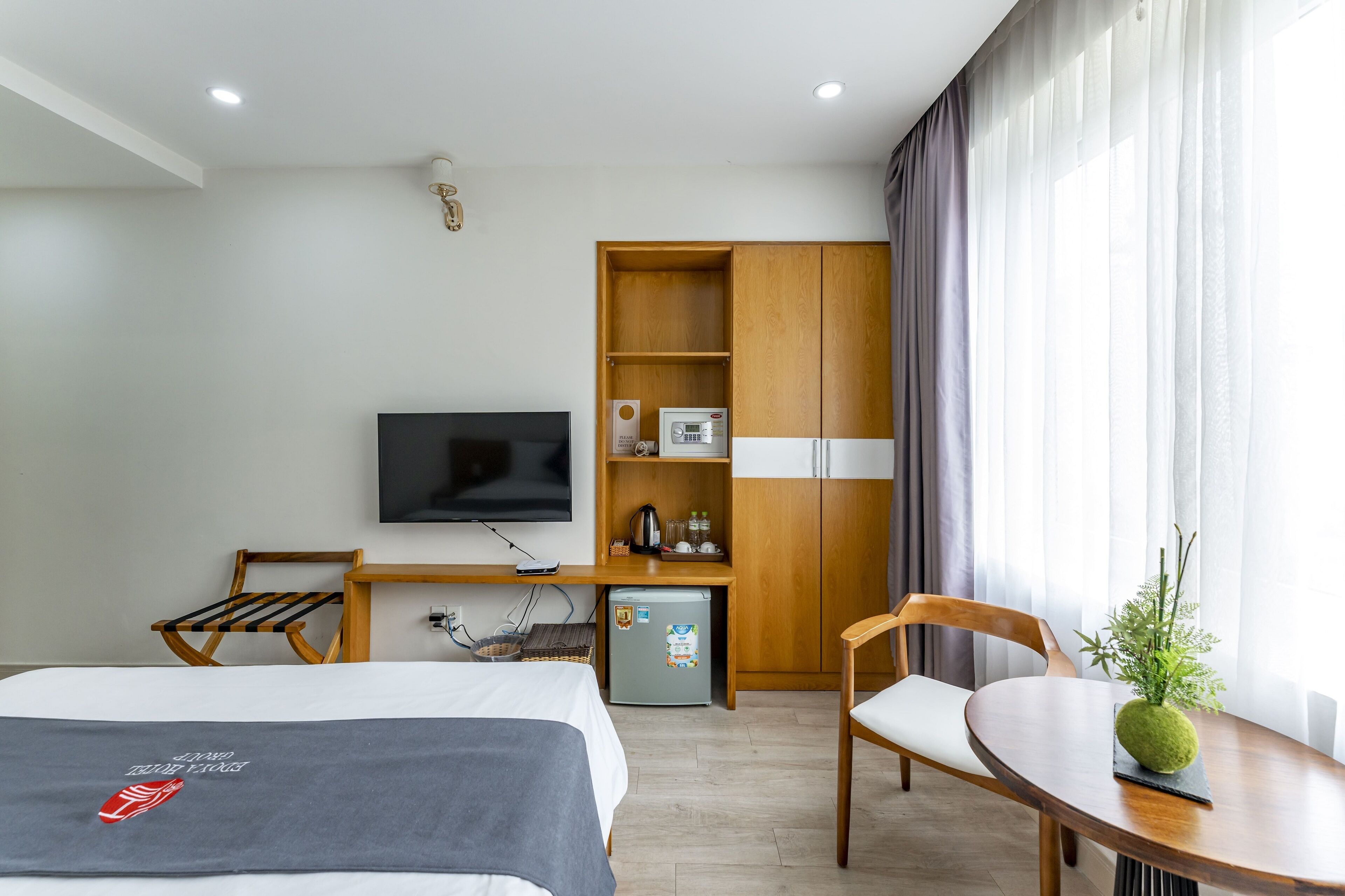 A hotel room with a bed, a desk with a TV, a small fridge, a wooden wardrobe, and a dining table with a plant.