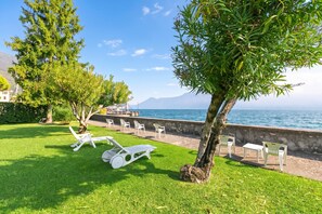Un área de estar al aire libre frente al mar con sillones reclinables y mesas, un césped y vistas al mar y montañas al fondo.