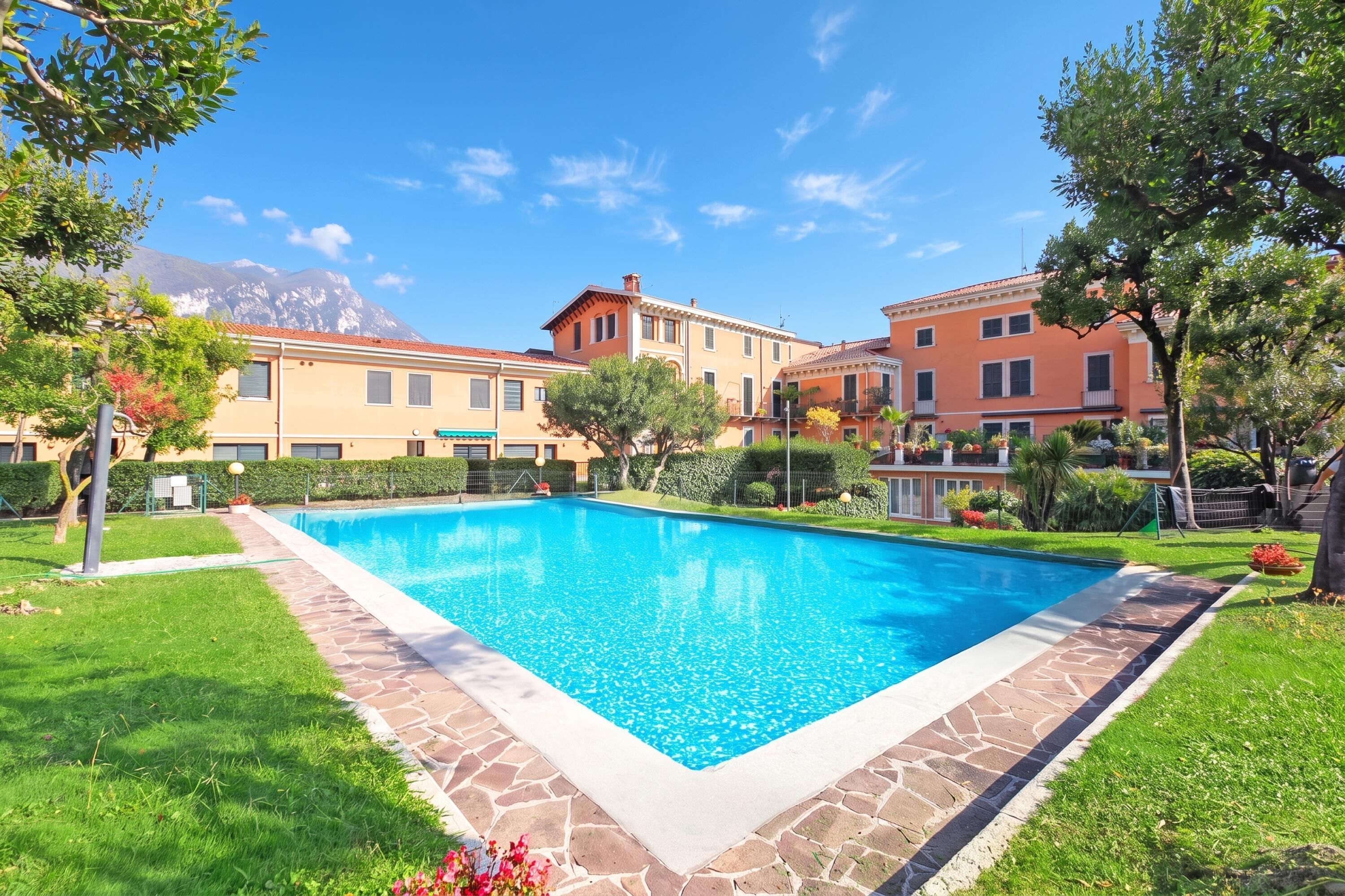 A swimming pool surrounded by a stone border, with a mountain in the background and a building with a red roof.