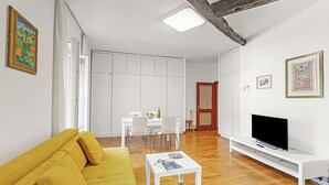 A living room with a yellow sofa, a white coffee table, and a TV on a white stand. There is a dining area with a table and chairs in the background. - Bogliaco Lake Front, Gargnano, Italy (Gargnano)