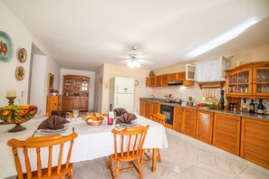 Comedor con una mesa dispuesta para cuatro, sillas de madera, un frutero y un refrigerador al fondo.