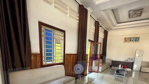 A living room with a glass-top coffee table, a beige sofa, and a white armchair. There is a window with blinds, a ceiling fan, and a radiator. - AHHA Residence Kampot (Kampot)