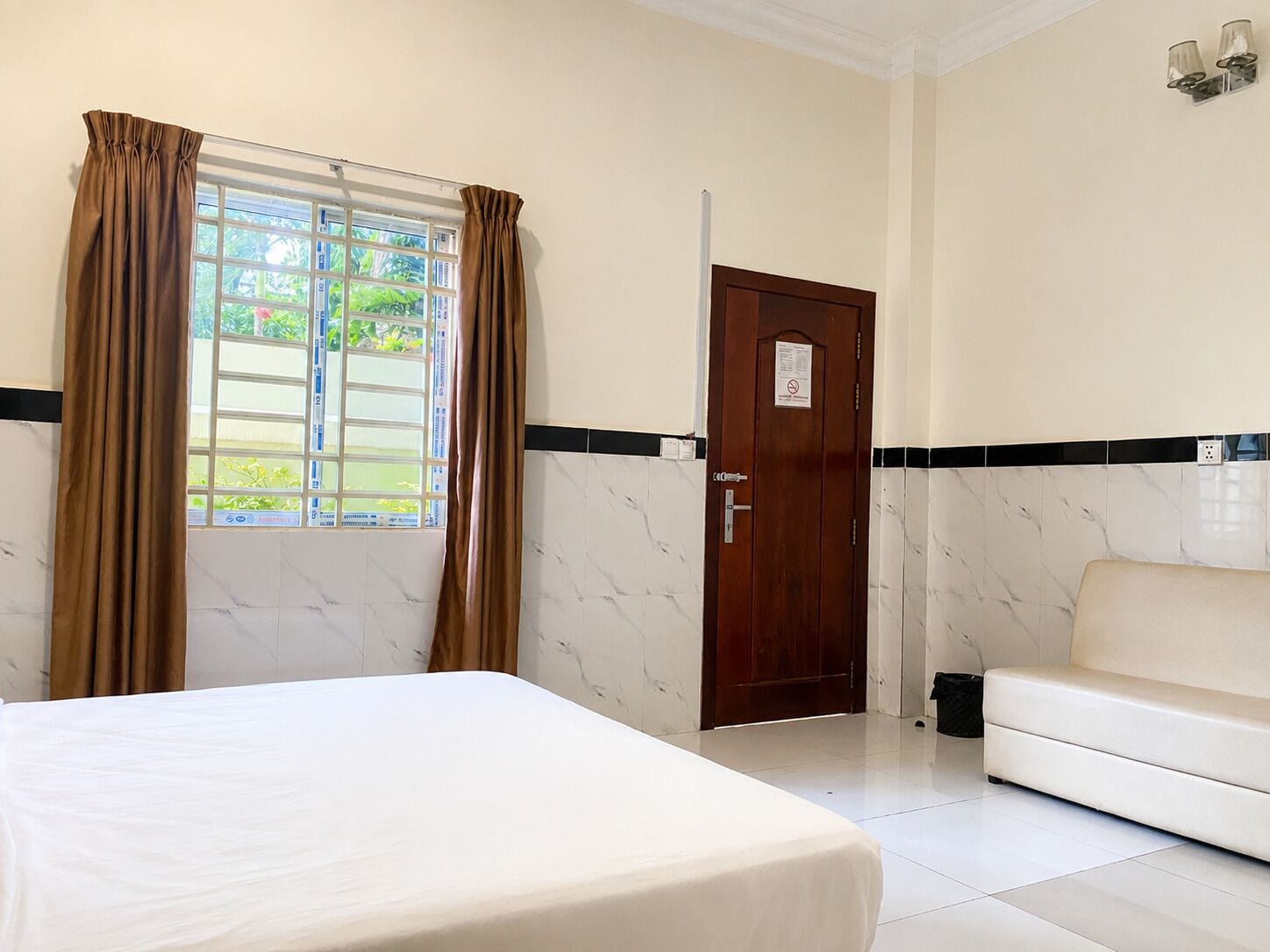 A hotel room with a bed, a sofa, a window with curtains, a door, and a wall-mounted light fixture.
