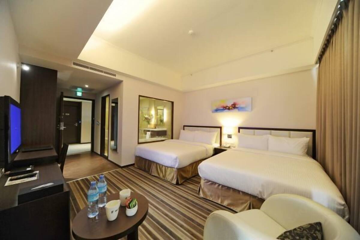A hotel room with two beds, a desk with a computer, a chair, a table with water bottles, and a painting on the wall.