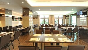 Un vaste espace de restauration dans l’hôtel, avec de nombreuses tables et chaises, de grandes fenêtres et un téléviseur à écran plat fixé au mur.