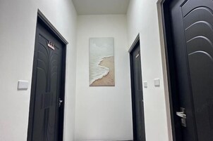 Un pasillo con dos puertas oscuras, una con una placa de número, y una obra enmarcada en la pared que representa una escena de playa.