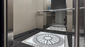 Un ascenseur à l’intérieur métallique, doté d’une barre de maintien et d’un sol orné d’un motif géométrique.