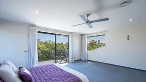 Et soveværelse med en stor seng, ventilator i loftet og en balkon med udsigt til træer.