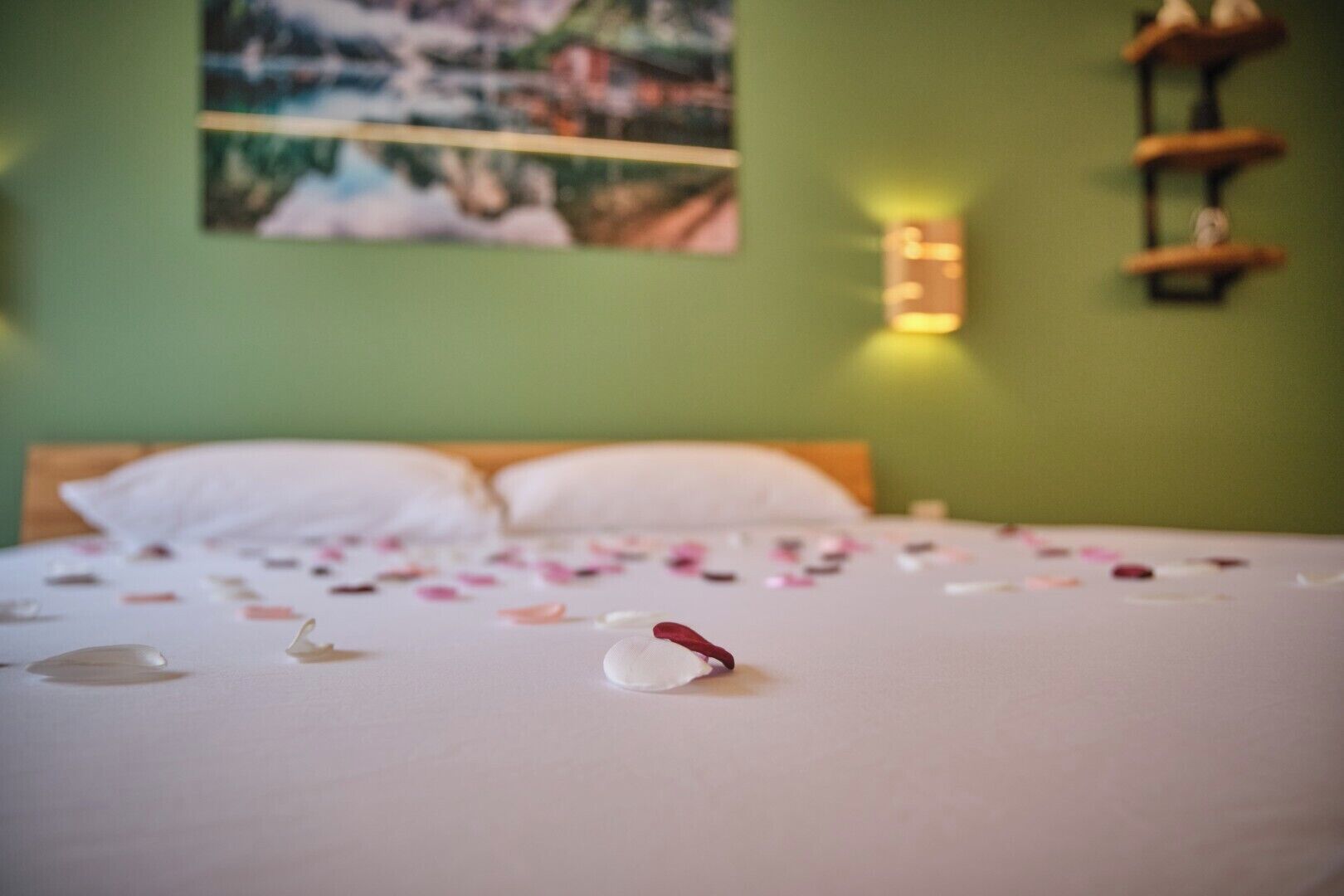 A bed with a white sheet and scattered petals, a painting on the wall, and a shelf with decorative items.