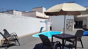 An outdoor patio with a pool, surrounded by chairs and an umbrella. - Cozy house in Caldas Novas with private heated pool. (Caldas Novas)