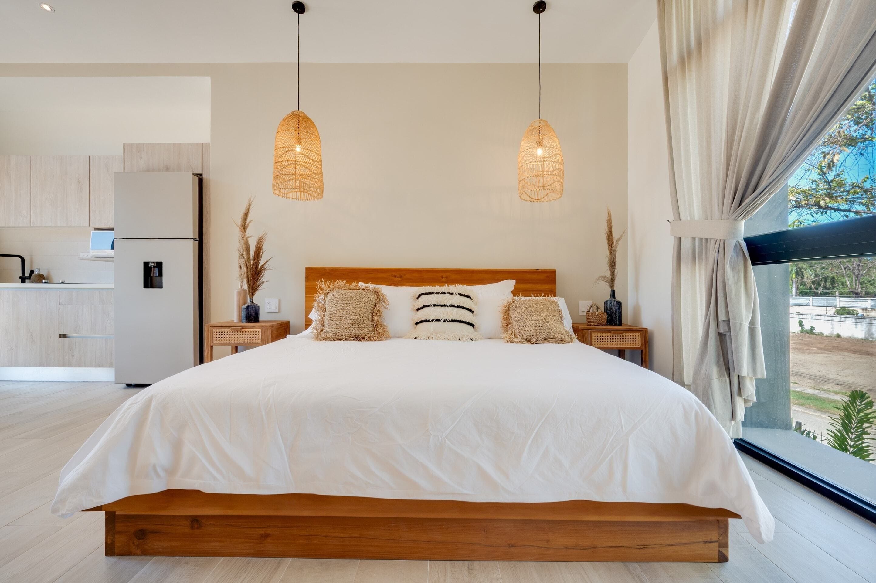 Ein modernes Schlafzimmer mit einem großen Bett, einem Holzkopfstück und Nachttischen. Links ist eine Küchenzeile erkennbar.