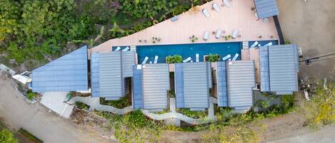 An aerial view of a hotel with a swimming pool, multiple rooms with blue roofs, and a parking area.