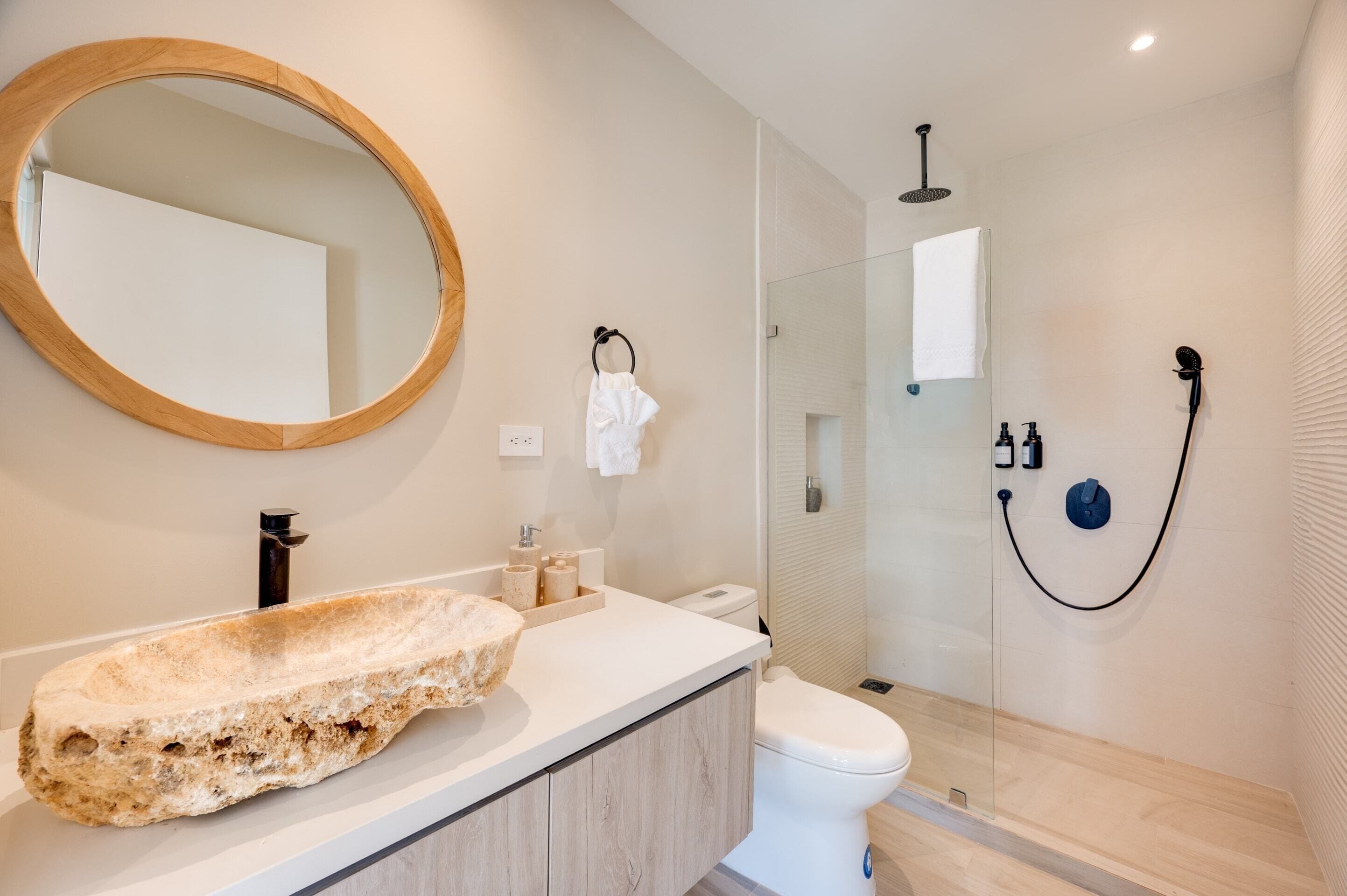 Ein modernes Badezimmer mit einem großen, ovalen Spiegel, einem Steinsink, einer Toilette und einer gläsernen Dusche.